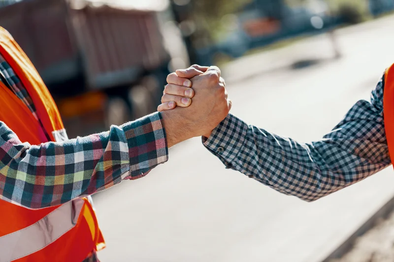 Zwei Menschen mit orangen Warnwesten und karierten Hemden geben sich einen freundschaftlichen Handschlag