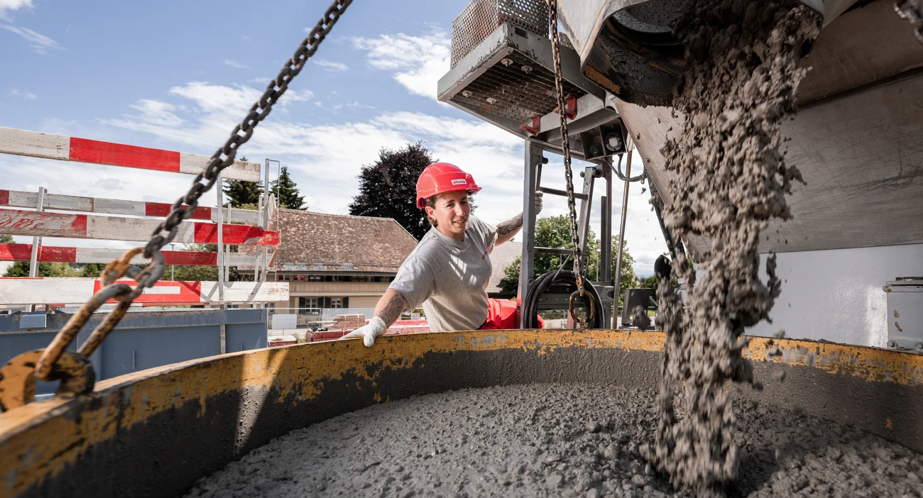 Eine Mitarbeiterin im grauen T-Shirt und orangem Helm lädt auf einer Baustelle Beton in einen Krankübel ab.
