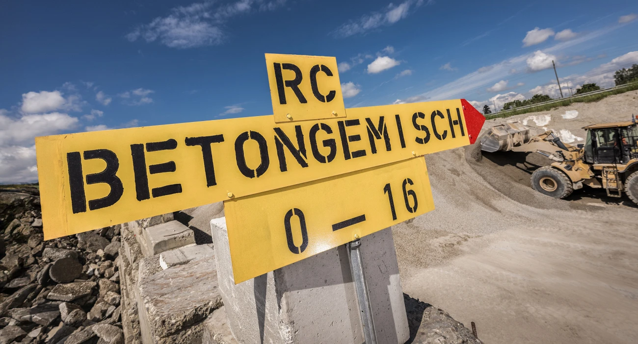 Ein grosses gelbes Schild mit der Aufschrift "RC-Betongemisch 0-16" vor der Kulisse des Depot- und Umschlagplatz in Mattstetten. Im Hintergrund ein gelber Pneulader, der Material auflädt.