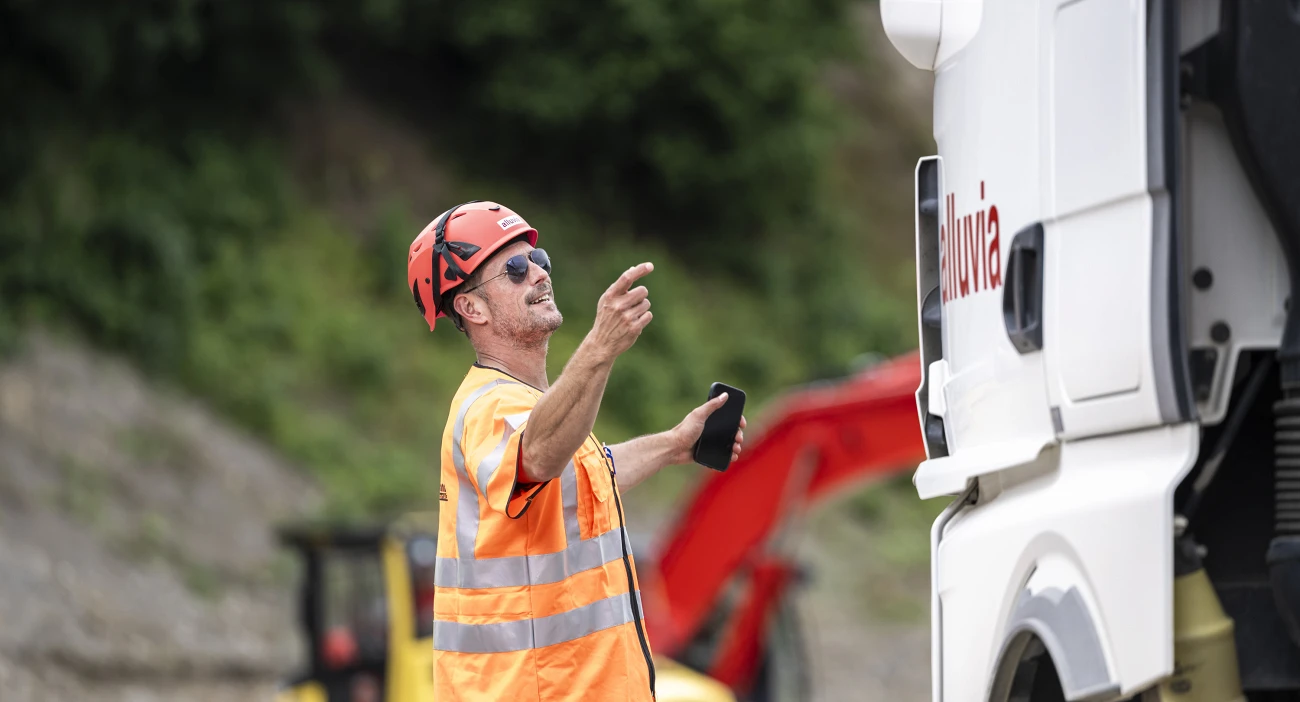 Ein Mitarbeiter mit rotem Helm und oranger Leuchtweste zeigt einem LKW-Fahrer den Weg auf die Baustelle. Eine weisse LKW-Kabine mit dem Schriftzug "alluvia".