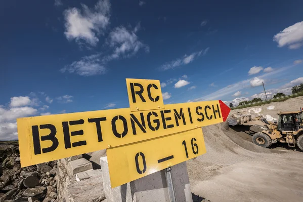 Ein grosses gelbes Schild mit der Aufschrift "RC-Betongemisch 0-16" vor der Kulisse des Depot- und Umschlagplatz in Mattstetten. Im Hintergrund ein gelber Pneulader, der Material auflädt.