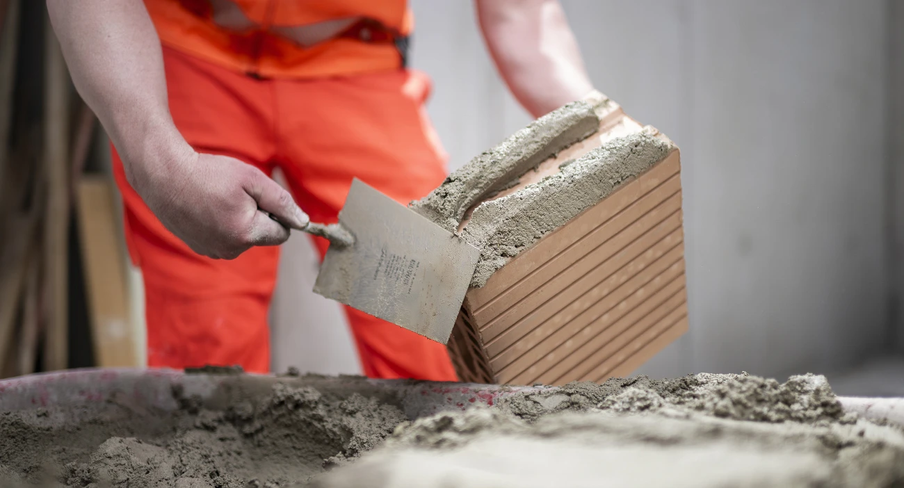 Ein Maurer in oranger Kleidung fügt mit einer Mörtelkelle Mörtel auf einen Ziegelstein.