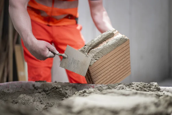 Ein Maurer in oranger Kleidung fügt mit einer Mörtelkelle Mörtel auf einen Ziegelstein.