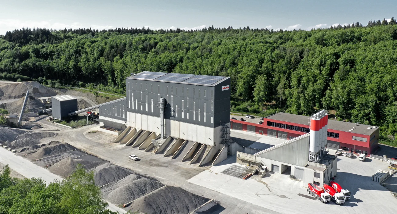 Kies- und Betonwerk Oberwangen mit rotem Betriebsgebäude und Wald im Hintergrund. Im Vordergrund drei rote LKWs der alluvia Flotte.