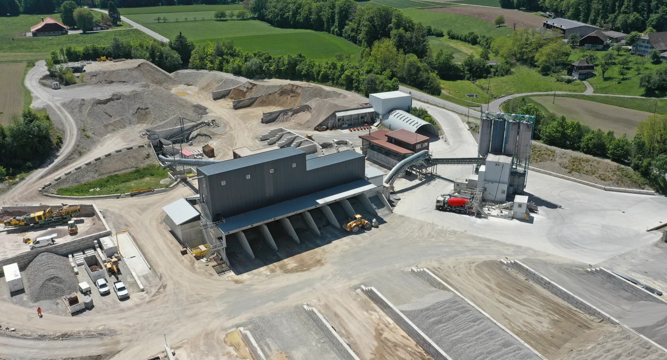 Drohnenansicht vom Kies- und Betonwerk in Berken. Im Zentrum das graue Kieswerk, im Vordergrund verschiedene Kiessilos und im Hintergrund Wiesen und Wald der Gemeinde Berken.