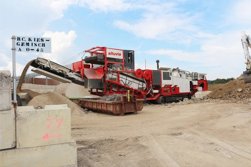 Eine mobiler Brecher im Einsatz im Kieswerk Berken, die recyceltes Kiesgemisch (RC Kies-Gemisch) sortiert. wird von einem Förderband gespeist. Im Hintergrund ist ein Schild mit der Aufschrift 'RC Kies-Gemisch A 0-45' zu sehen.